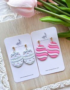 two pairs of pink and white earrings sitting on top of a wooden table next to flowers