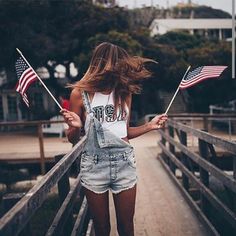 a woman walking across a bridge holding two american flags in one hand and an american flag in the other