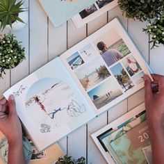 two hands are holding an open book with photos on it and some plants in the background