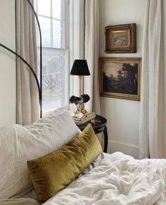 a bed with white sheets and pillows in front of a painting on the wall next to a window