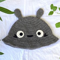 a crocheted hat with eyes and ears on top of a white sheet next to green leaves