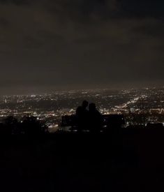 two people are sitting on a bench at night overlooking the city lights in the distance