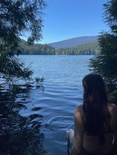 a woman sitting on the edge of a body of water with trees in the background