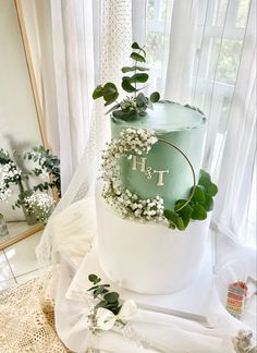 a three tiered cake with greenery and flowers on top, sitting in front of a window