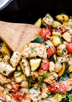 a skillet filled with chicken, zucchini, and tomato salad next to a wooden spoon