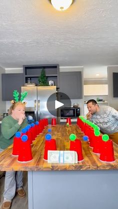 two men and a woman are in the kitchen playing with plastic cups on a wooden table