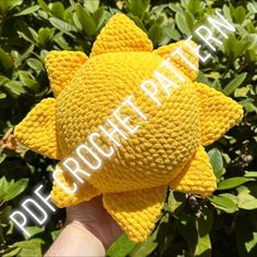 a hand holding up a yellow crocheted sunflower in front of some bushes