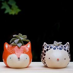 two ceramic pots with plants in them on a table next to each other, one has an animal face and the other has a cat's head