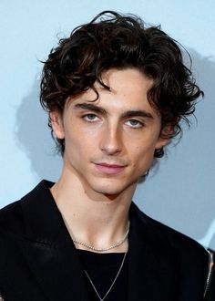 a young man with curly hair wearing a black shirt and silver necklace, looking at the camera