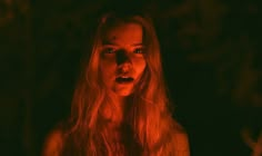 a woman with long blonde hair standing in the dark holding a lit candle and looking at the camera