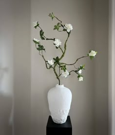 a white vase with flowers in it sitting on a black stand next to a wall