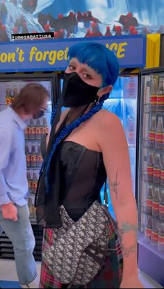 a woman with blue hair wearing a black face mask and standing in front of a vending machine