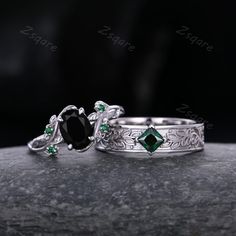 two wedding rings with black diamonds and green emeralds on the sides, sitting on top of a rock