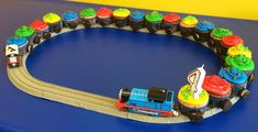 a toy train set with cupcakes on the track in front of it that is shaped like a birthday cake