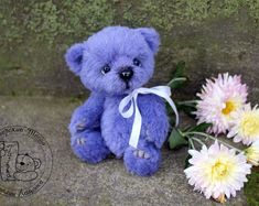 a purple teddy bear sitting next to some flowers on the ground with moss growing behind it