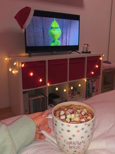 a person sitting on a bed with a cup of hot chocolate and marshmallows in front of a television