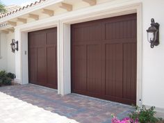 two brown garage doors in front of a white house with pink flowers on the sidewalk