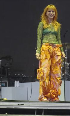 a woman standing on top of a stage with her hands in her pockets and wearing yellow pants