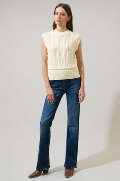 a woman standing in front of a white background wearing jeans and a sweater with cables on it