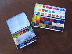 two tins of watercolor paint sitting on top of a wooden table next to each other