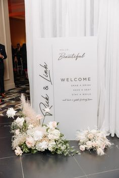 a welcome sign with flowers and greenery on the floor next to a white backdrop