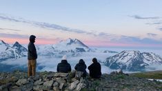 three people sitting on top of a mountain looking at the clouds and mountains in the distance