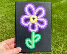 a hand holding up a black square with purple and green flowers on the outside, in front of some grass