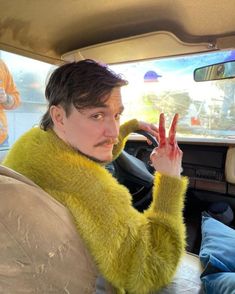 a person sitting in a car making the peace sign with their hand and wearing a fuzzy yellow sweater