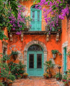 an orange building with green doors and purple flowers on the windows sill, surrounded by potted plants