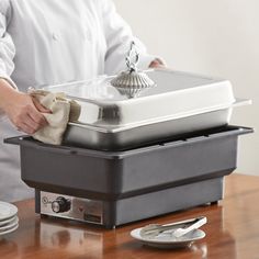 a person is holding a cloth in front of a food warmer on a table with plates and utensils