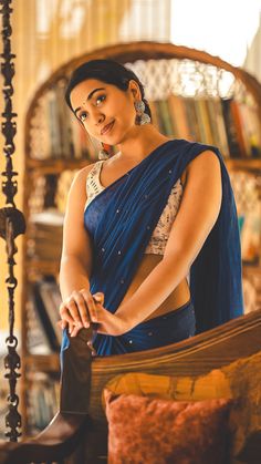 a woman in a blue sari is standing near a book shelf and looking at the camera