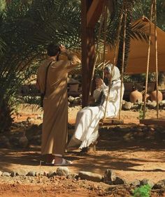 two people dressed in traditional garb sitting on swings and talking to each other, with palm trees in the background