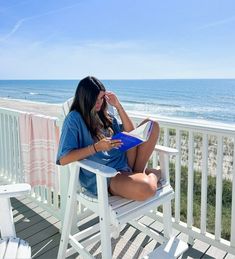 a woman sitting in a chair on top of a balcony next to the ocean reading a book