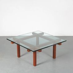 a square glass table with wooden legs on a gray floor in front of a white wall