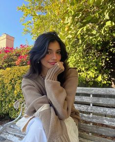 a woman sitting on top of a wooden bench next to trees and bushes with her hand under her chin