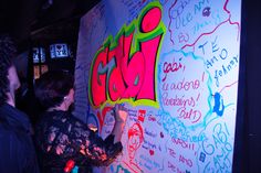 two people writing on a large wall with colorful chalk and neon lights in the background