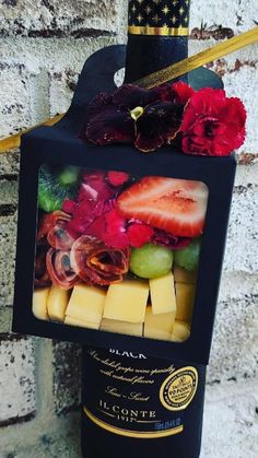 a wine bottle wrapped in black paper with fruit and flowers on the top, sitting next to a brick wall