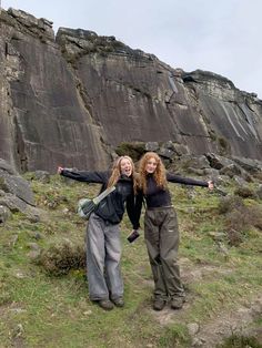 two young women standing on top of a grass covered field next to a large rock formation