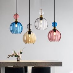 four different colored glass pendant lights hanging over a table with two chairs and a vase on it