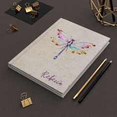 an open notebook with a dragonfly on it next to some gold and black items