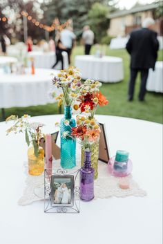 colorful vases filled with flowers sit on top of a white table cloth at an outdoor wedding