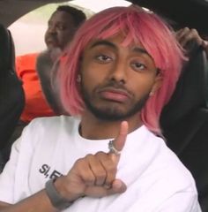 a man with pink hair sitting in a car and pointing to the side while wearing a white t - shirt