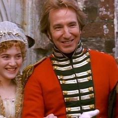 a man and woman dressed in period costumes smiling at the camera, with one wearing a red coat