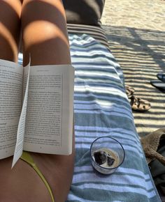 a person laying on the beach with their legs crossed reading a book next to a glass of water