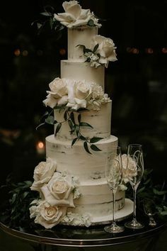 a three tiered cake with white flowers and greenery on the top is surrounded by wine glasses