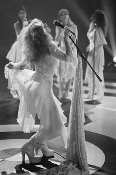 a woman in white dress standing on stage with microphone and other women behind her wearing long dresses