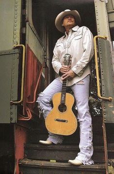 a man with a guitar standing on the steps of a train car, wearing white jeans and a cowboy hat