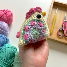 a hand holding a small crocheted bird in front of some yarn and scissors