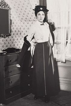 an old black and white photo of a woman standing in front of a dresser with a hat on her head