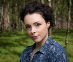 a woman standing in the woods wearing a blue shirt and looking at the camera with a smile on her face